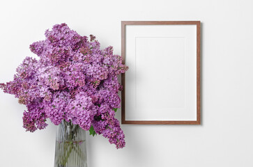 Blank wooden picture frame mockup with lilac flowers bouquet over white wall, mockup with copy space