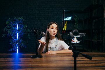 Backstage photo of female blogger sitting with microphone in cozy studio with blue light and...