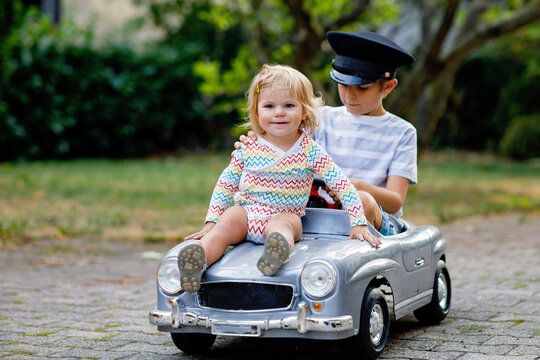 Two Happy Children Playing With Big Old Toy Car In Summer Garden, Outdoors. Kid Boy Driving Car With Little Toddler Girl Inside. Laughing And Smiling Kids. Family, Childhood, Lifestyle Concept