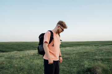 A stylish young man with a beard and wearing sunglasses with a backpack on his back is walking through an endless field against the background of a blue sky at sunset