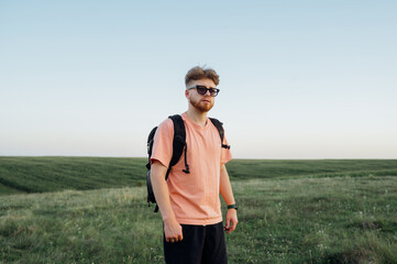 Portrait of a handsome bearded young man in stylish casual clothes and with a backpack on his back in an endless field against a clear blue sky at sunset