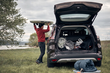 Windsurfer and camper unpacking equipment from a car in nature near the lake shore.
