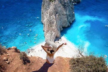 A tourist woman sits at the viewpoint of Keri and enjoys the view of the famous Mizithres rocks with turquoise sea at Zakynthos island, Greece - obrazy, fototapety, plakaty