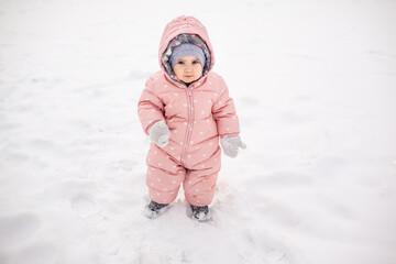 Fototapeta na wymiar A todler walks in the snow. A one year old baby dressed a warm pink jumpsuit, a winter hat and mittens