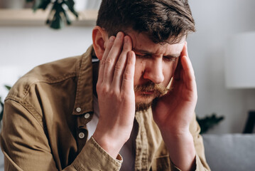 Close-up of upset exhausted young bearded man 30s sitting on sofa in living room at home, feels unwell, suffering migraine, fatigue, experiences of personal troubles, feeling of anxiety concept