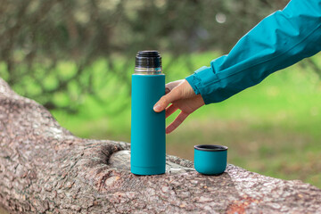 a human hand holds a steel green thermos, a mug with hot tea nearby on a branch of an old coniferous tree in the forest. Nature, picnic, camping