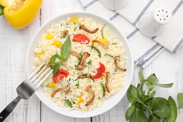 Bowl of delicious couscous with vegetables and basil served on white wooden table, flat lay