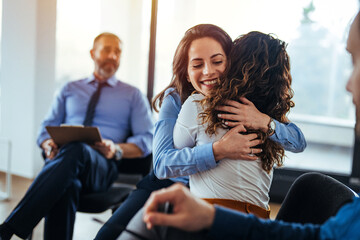 Young adult woman embracing and supporting friend during support group therapy session with diverse women. Two women hug in therapy session. Group therapy session, empathy concept