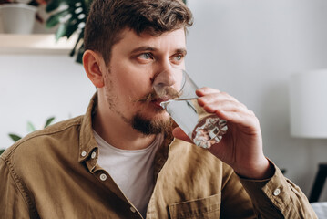 Close up of young bearded man drinking fresh water from glass, sitting on sofa in living room at...