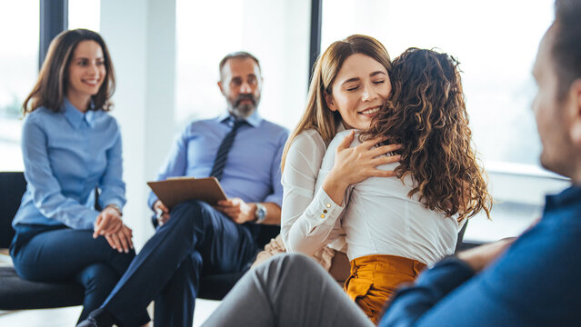 Young adult woman embracing and supporting friend during support group therapy session with diverse women. Two women hug in therapy session. Group therapy session, empathy concept