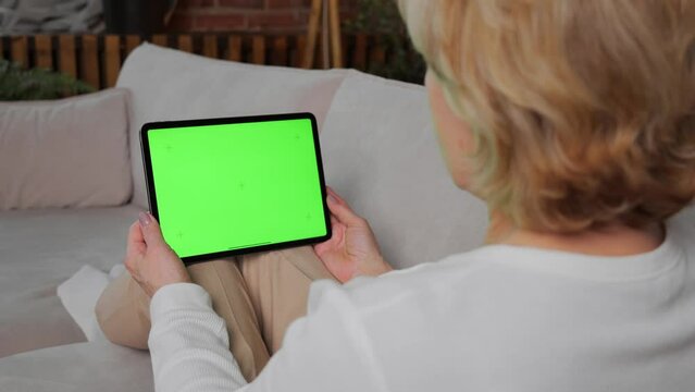 An elderly woman of 60s is sitting at home on a pleasant sofa with a graphic tablet in her hands, which has a green screen turned on to integrate media images of a program or video. 4k footage