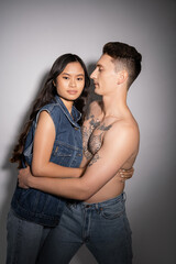 brunette asian woman in denim vest looking at camera near muscular tattooed man on grey background.