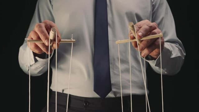 A businessman in a gray shirt and black tie controls a puppet with a wooden manipulator and strings. The puppeteer manipulates the puppet by pulling the ropes with both hands. Close up.