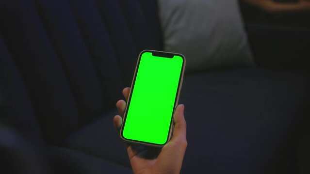 Footage of unrecognizable female hand holding smartphone with green screen in evening. Woman in plaid shirt touching screen of gadget, scrolling on background of blue sofa indoors