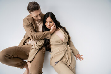 high angle view of interracial romantic couple in beige suits touching each other while posing on grey background.