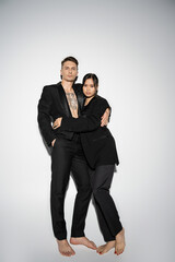 full length of barefoot interracial couple in black elegant outfit embracing and looking at camera on grey background.