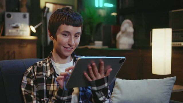 Footage of friendly-looking young Caucasian woman with nose piercing resting on sofa with digital tablet. Smiling girl in comfortable casual clothes enjoying leisure time at home indoors. Technology