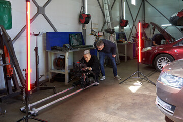 Fototapeta na wymiar A cameraman and a director are setting up a professional video camera on a film set with cars and red light fixtures. camera on operator platform and rails