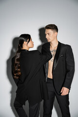 long haired asian woman and tattooed man in black elegant attire looking at each other on grey background.