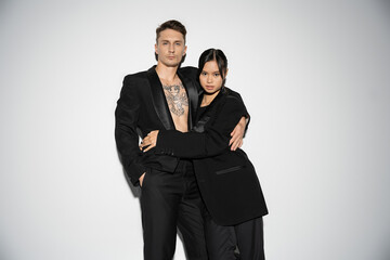 fashionable multiethnic couple in black elegant suits embracing and looking at camera on grey background.