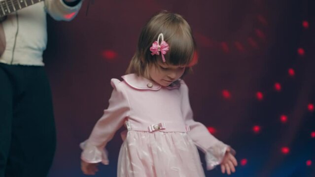 Little girl on stage in pink vintage dress, she dances, her father plays acoustic guitar. Color music is shining. Performance on stage. Early development of children