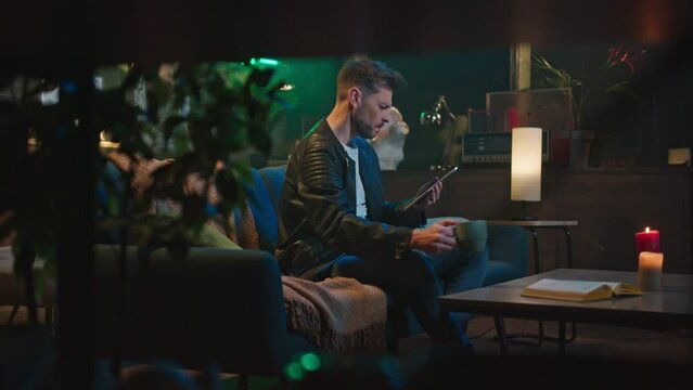 Horizontal shooting of handsome focused young Caucasian man sitting on sofa, drinking tea and using tablet in evening. Beautiful cozy interior of apartment. Indoors