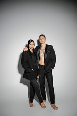 full length of tattooed man and asian woman in black fashionable outfit looking at camera on grey background.