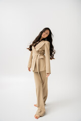 full length of barefoot woman with long hair posing in fashionable pantsuit on grey background.