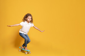 Full length of little girl in white polo, blue jeans who skating on penny board on yellow background