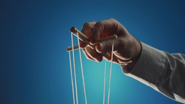 The hand of a man in a gray shirt controls a puppet using a wooden manipulator and strings on a blue background. Hand handling at puppet by pulling strings to make the character move. Close up.