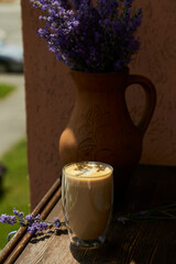 Glass with a double bottom of latte on wooden table under hard shadows. Lavender flowers decorations outside in the cafe terrace. Atmospheric coffee break.