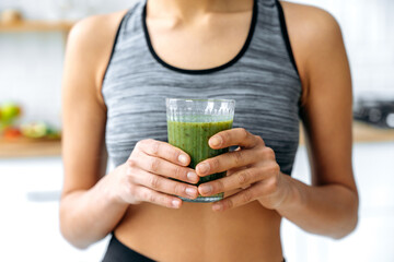 Close-up photo of a green fresh smoothie in a glass cup in female hands. Sporty woman prepared...