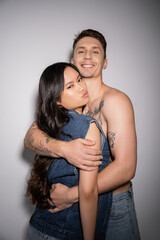 cheerful tattooed man embracing stylish asian woman pouting lips on grey background.