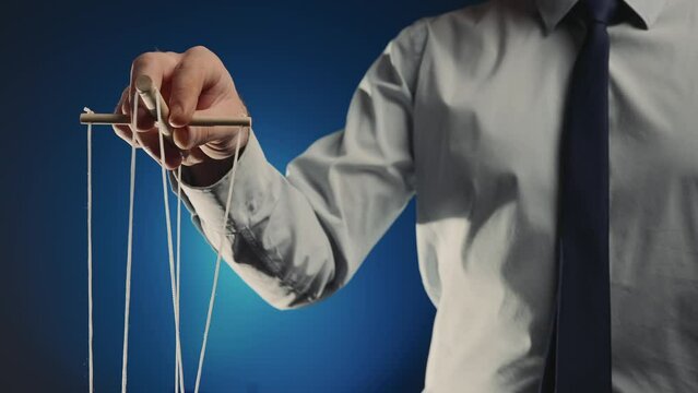 A businessman in a gray shirt and black tie controls a puppet with a wooden manipulator and strings. Hand handling at puppet by pulling strings to make the character move. Blue background. Close up.