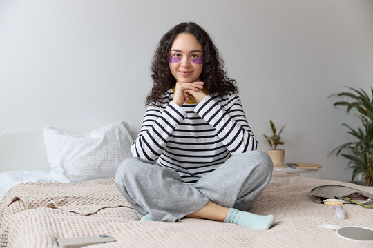 A curly adult woman with long brown hair smiles while sitting on the bed during a beauty photo shoot. She wears casual, striped clothing and looks directly at the camera. Sits in a relaxed position.