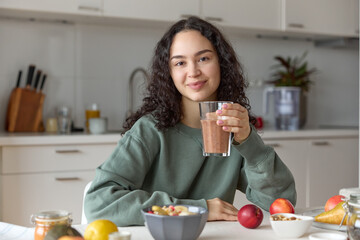 Close up of a young curly haired smiling woman vegan holding a glass of smoothie enjoying having a...