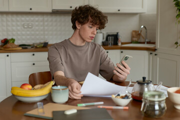 A young curly haired handsome man wearing a beige t-shirt sitting at a kitchen table surrounded by comforts of domestic life pays the bills working reviewing papers while having breakfast in kitchen.