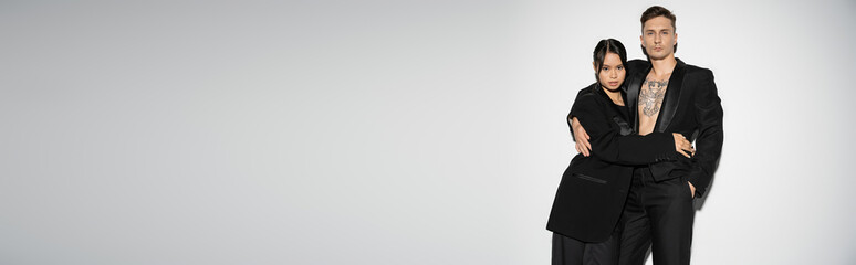 brunette asian woman and tattooed man wearing black stylish suits and embracing on grey background, banner.