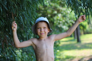 A cute boy with a bare torso is holding on to tree branches in the park. Summer vacation.