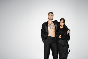tattooed man in black trendy suit embracing elegant asian woman while posing with hand in pocket on grey background.