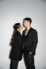 side view of smiling asian woman touching tattooed chest of man in black suit on grey background.