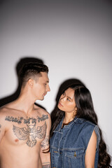 shirtless man with tattooed body and brunette asian woman with long hair looking at each other on grey background.