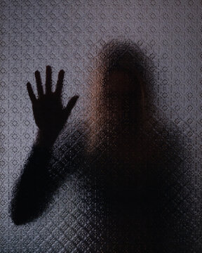 Blurred silhouette of a mysterious woman standing behind frosted glass with hand against the glass. Concept of isolation, loneliness or depression
