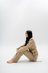side view of young and barefoot asian woman in beige pantsuit sitting on grey background.