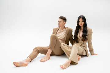 full length of barefoot interracial couple in beige suits sitting on grey background.