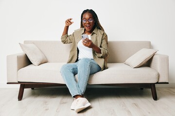 Plakat African American woman business freelancer working sitting on the couch at home in the phone, business calls and messages happiness smile, home clothes and eyeglasses, light interior background.