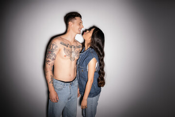 long haired asian woman in denim outfit sticking out tongue near smiling tattooed man on grey background.