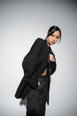 brunette asian woman in black elegant blazer and trousers standing with hand in pocket on grey background.