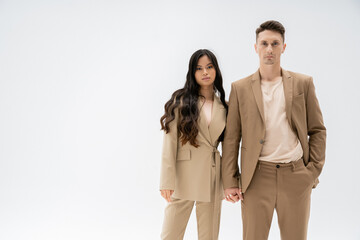 interracial couple in fashionable outfit holding hands and looking at camera isolated on grey.