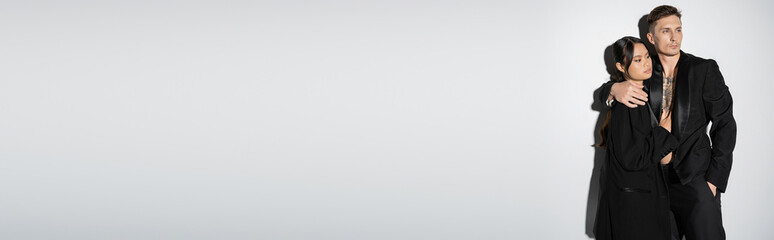 interracial couple in black fashionable outfit looking away on grey background, banner.